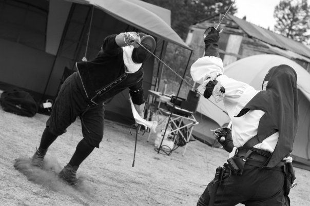Author in black, posed with sword held high, threatening another fencer who is defending with off-hand dagger