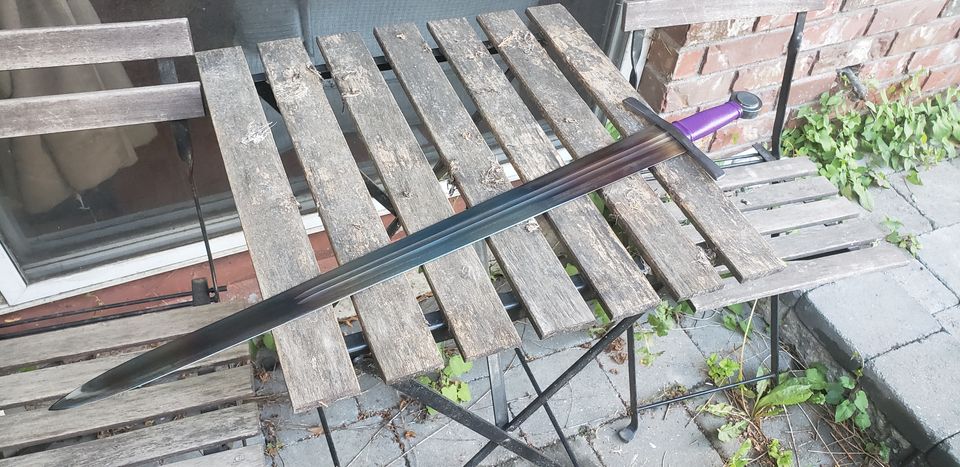 Sharp medieval sword with rainbow-coloured blade and purple handle-wrap, displayed on an outdoor table