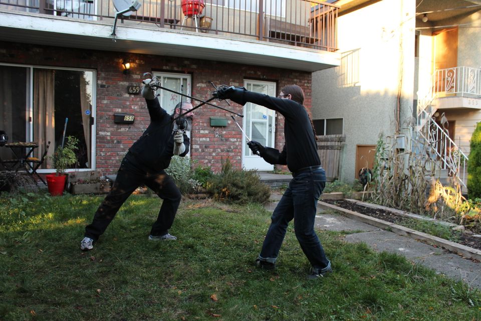 Two folks sparring with rapiers and daggers, on the lawn in front of a house